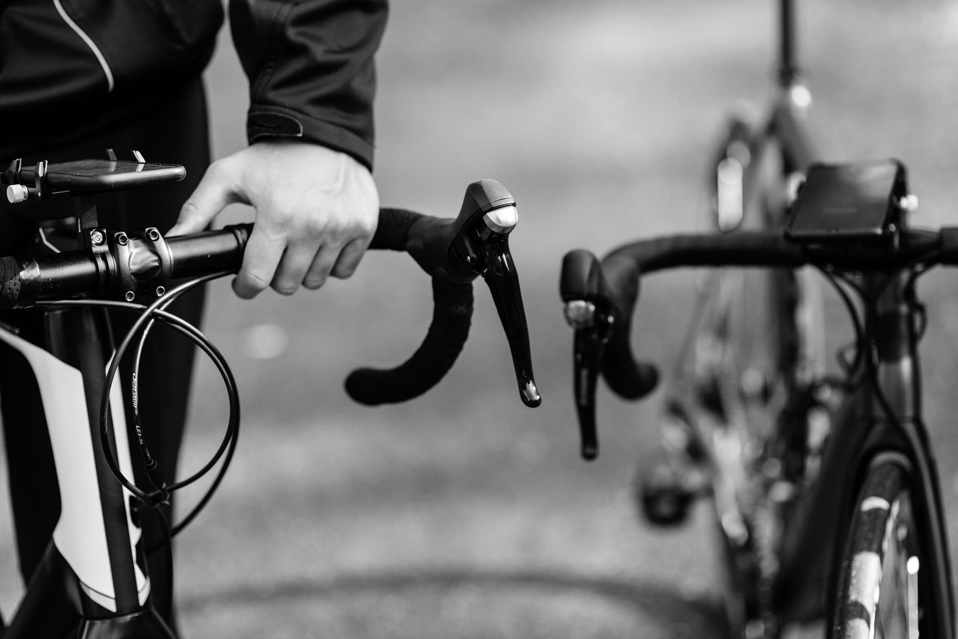A cyclist's gloved hands resting on the brake hoods of a road bike handlebar in bright light.