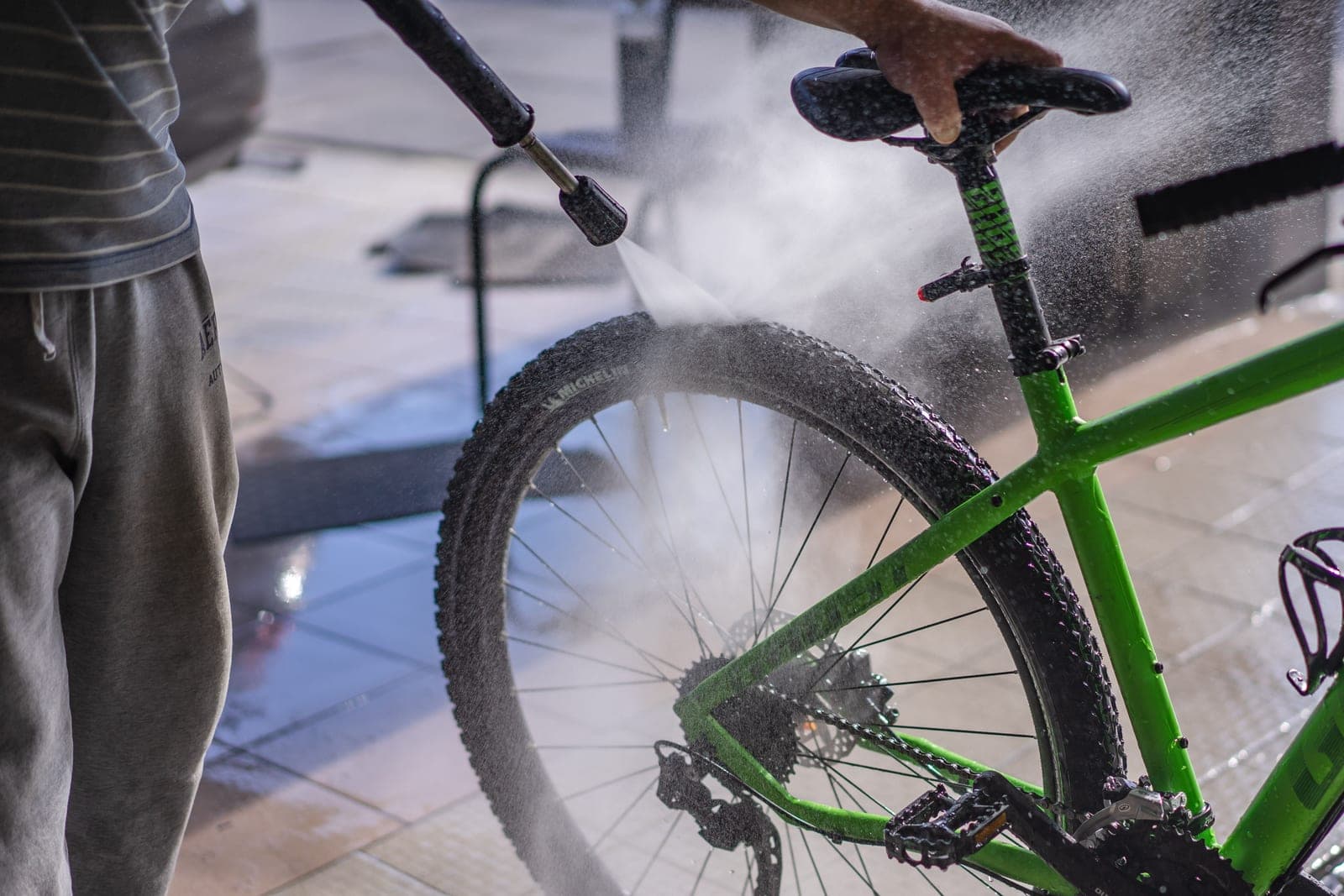 A person washing a green bicycle outdoors with a hose, focused on the wheel and frame.